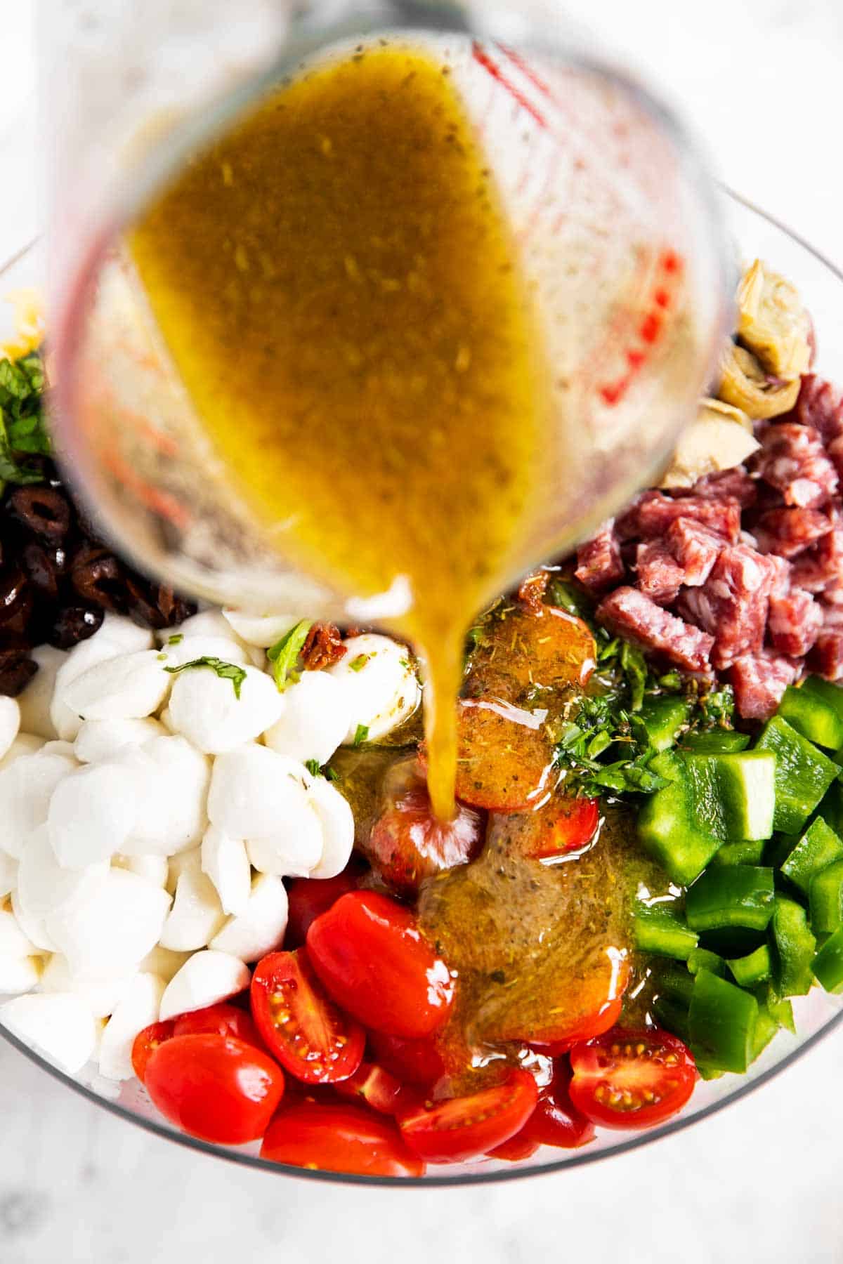 Italian dressing pouring from glass measuring jug over Italian pasta salad ingredients in glass bowl