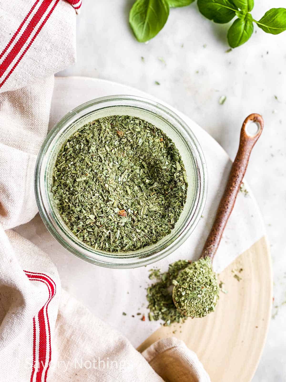 Italian seasoning in small glass jar on serving platter, with ceramic spoon next to it