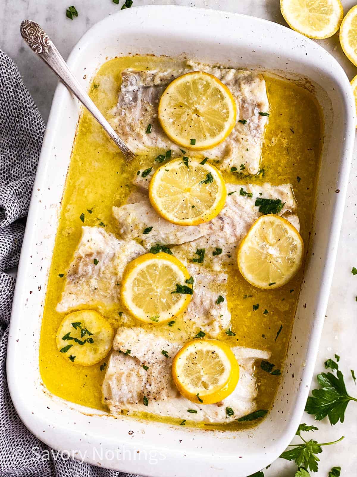 overhead view of white casserole dish with four cod pieces in lemon garlic butter sauce, topped with lemon slices and chopped parsley