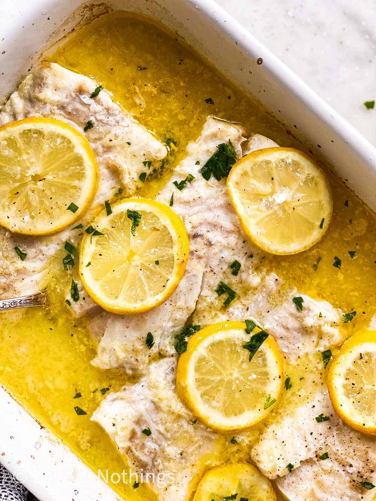 overhead view of four cod loin fillets in white casserole dish with lemon sauce and lemon slices