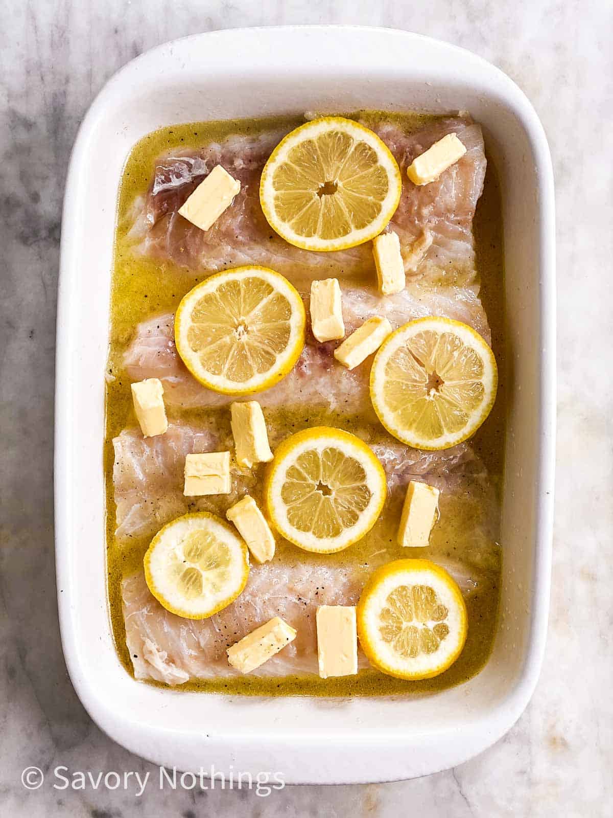 unbaked cod loin fillets in white casserole dish, topped with lemon slices and butter pieces