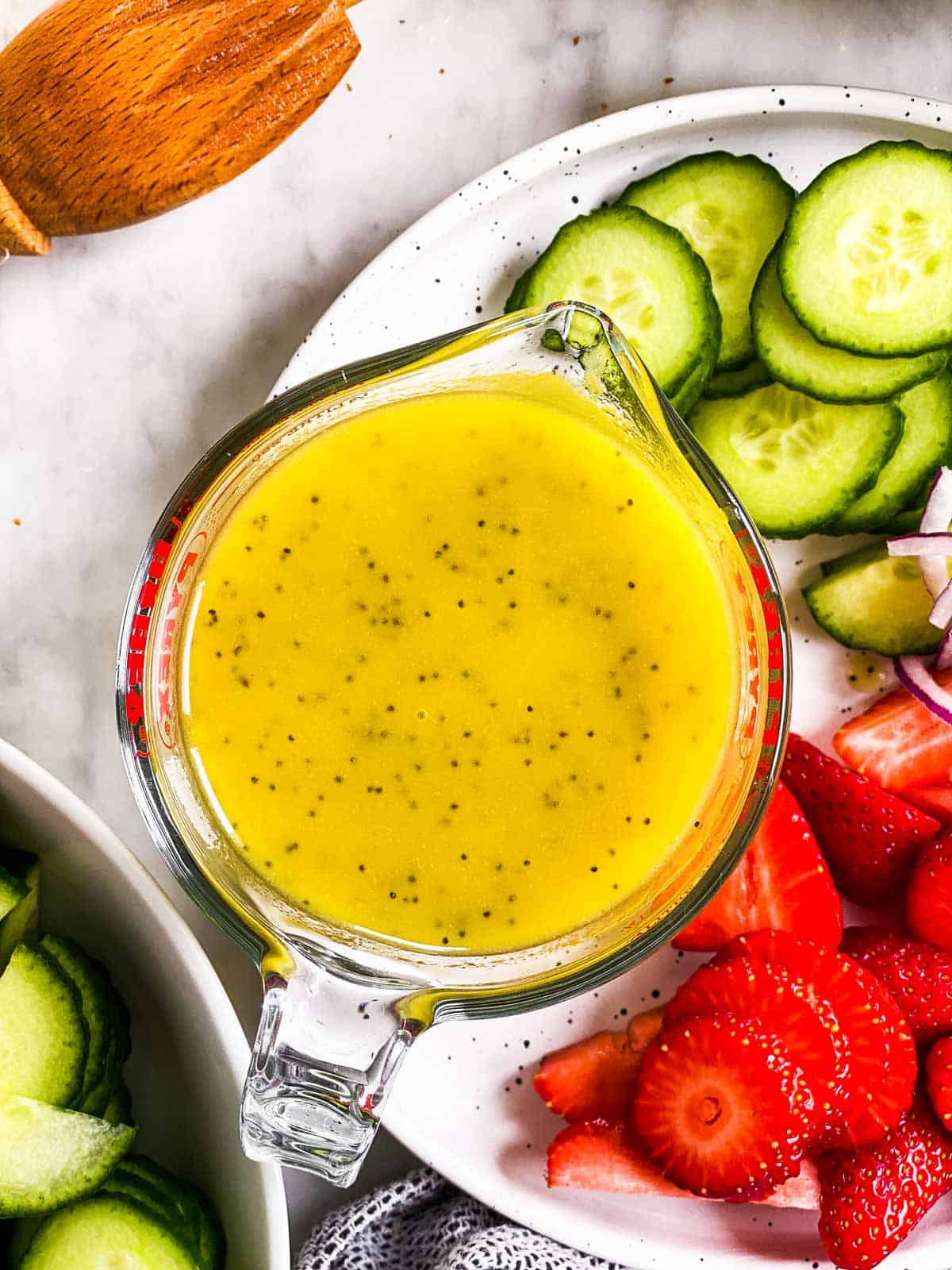 overhead view of lemon poppy seed salad dressing in glass measuring jug on plate with strawberries and cucumbers