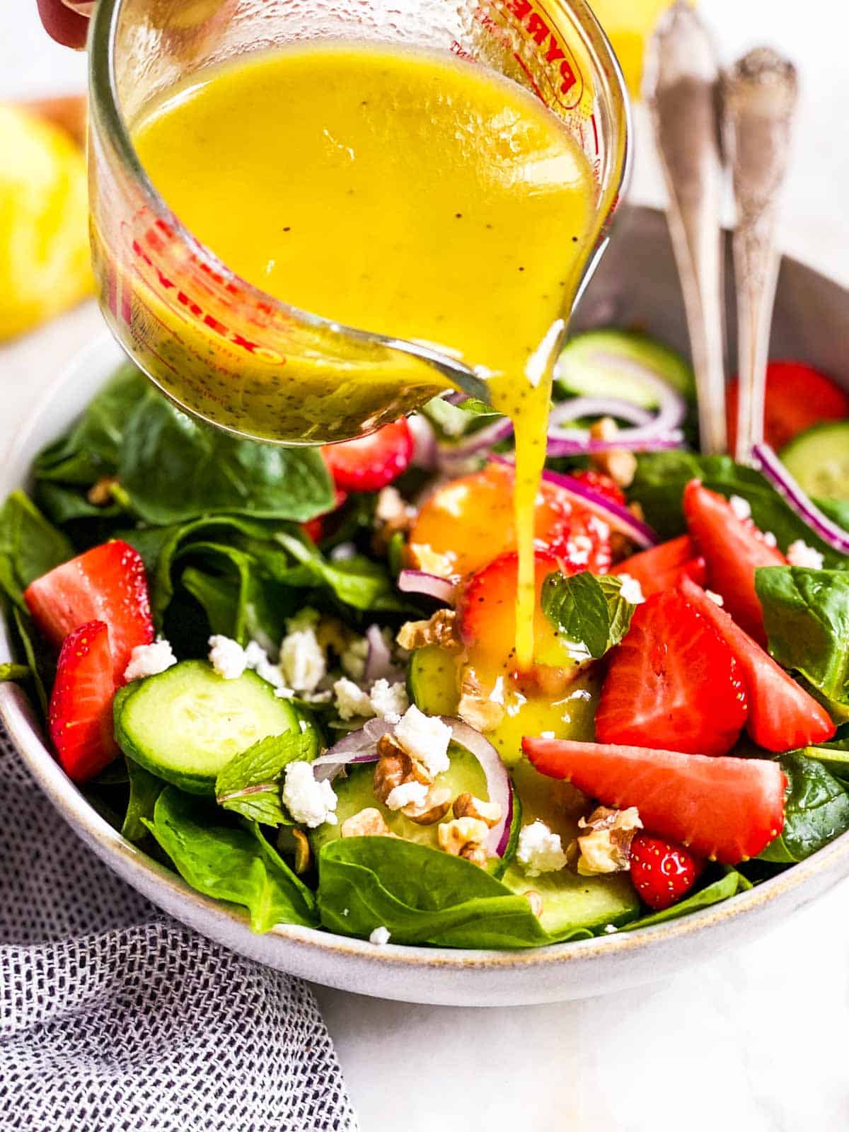 lemon poppy seed salad dressing pouring over spinach salad from glass measuring jug