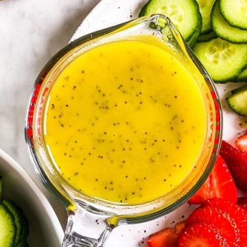 overhead view of lemon poppy seed salad dressing in glass measuring jug on plate with strawberries and cucumbers