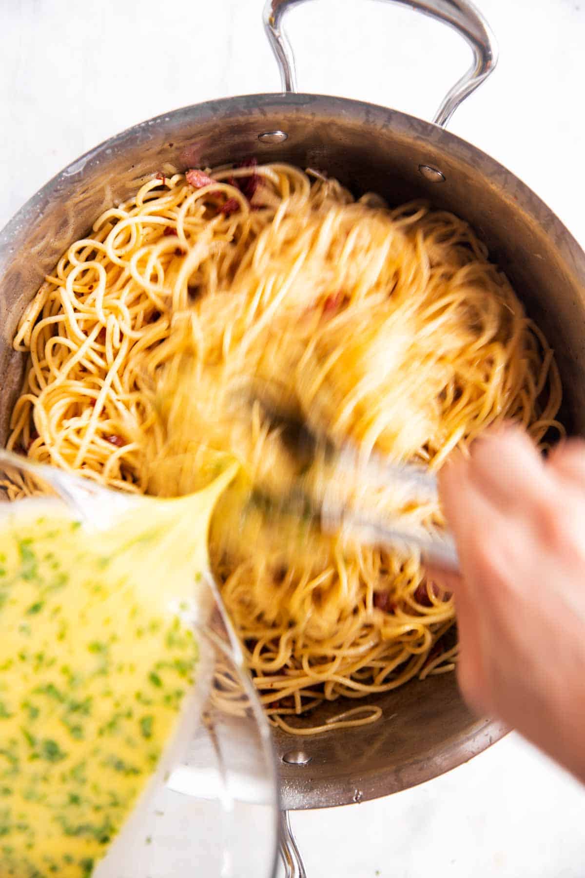 female hand tossing spaghetti with beaten eggs in stainless steel skillet