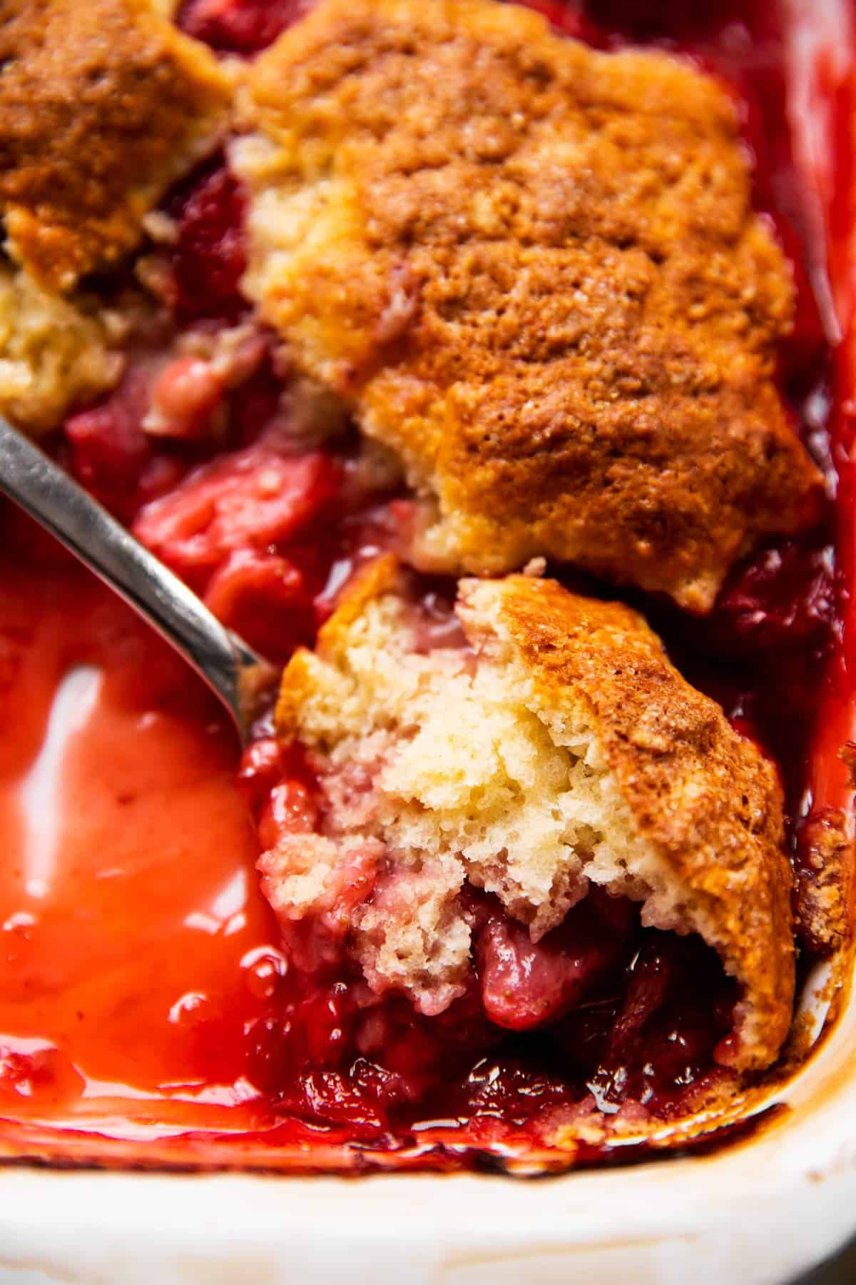 close up photo of spoon scooping strawberry cobbler from white casserole dish