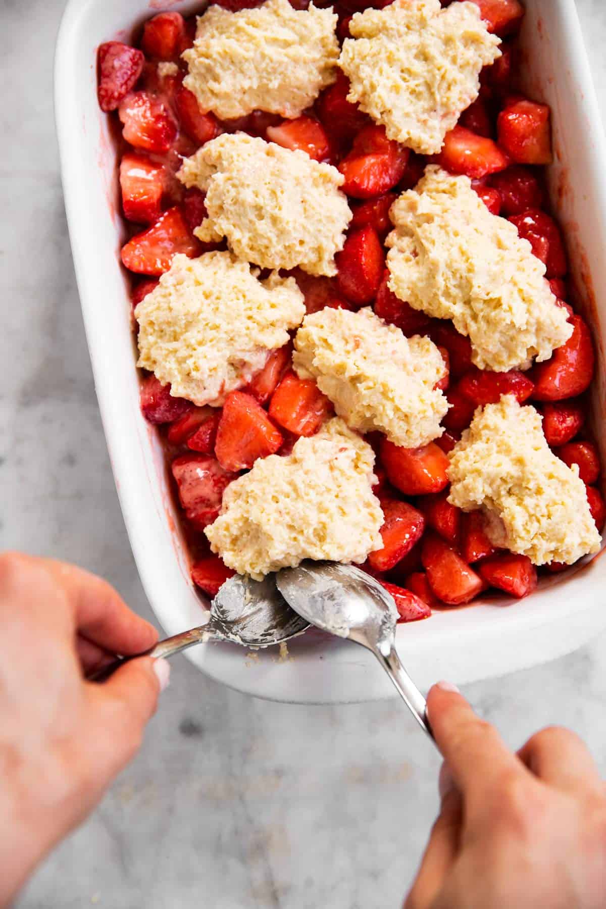 female hands placing drop biscuits on top of strawberry filling in white casserole dish