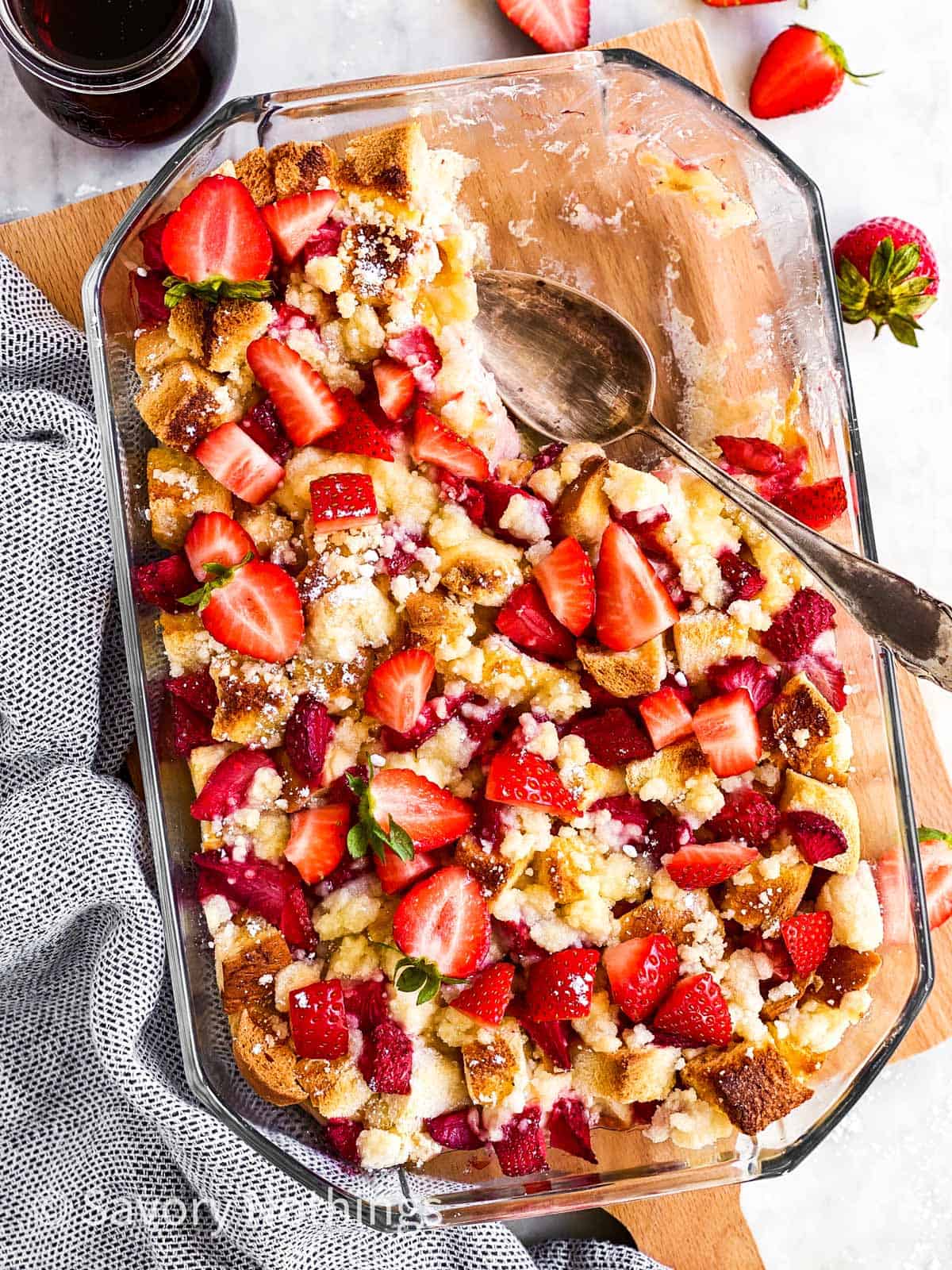 overhead view of baked strawberry French toast casserole in glass dish with a scoop taken out, sitting on wooden chopping board