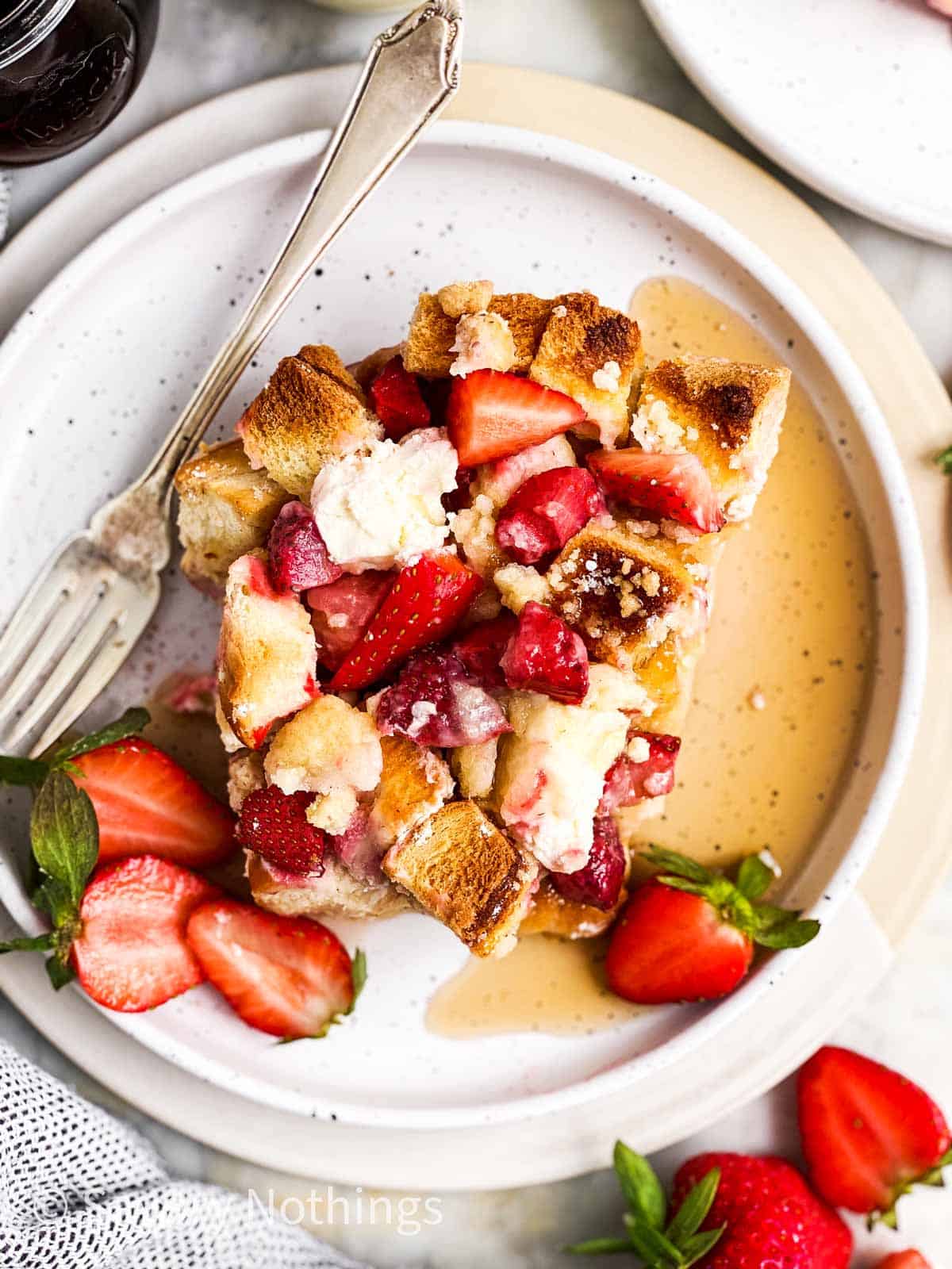 overhead view of a slice of strawberry French toast casserole on a white plate with maple syrup