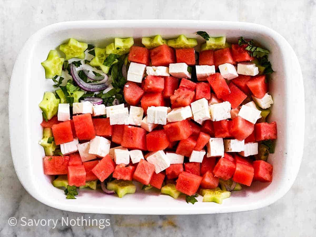 watermelon and feta cheese arranged in stripes over cucumber stars, herbs and red onion in rectangular white dish