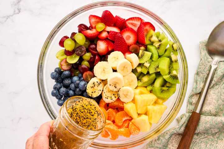 Pouring dressing over cut up fruit in a glass bowl.