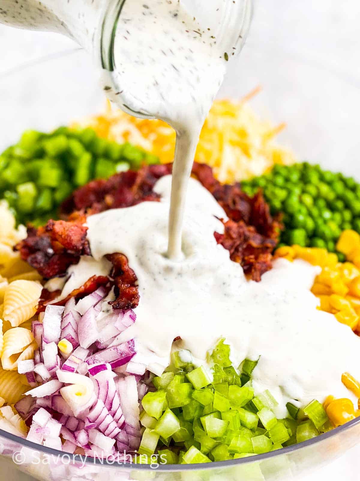 ranch dressing pouring over pasta salad ingredients in glass bowl