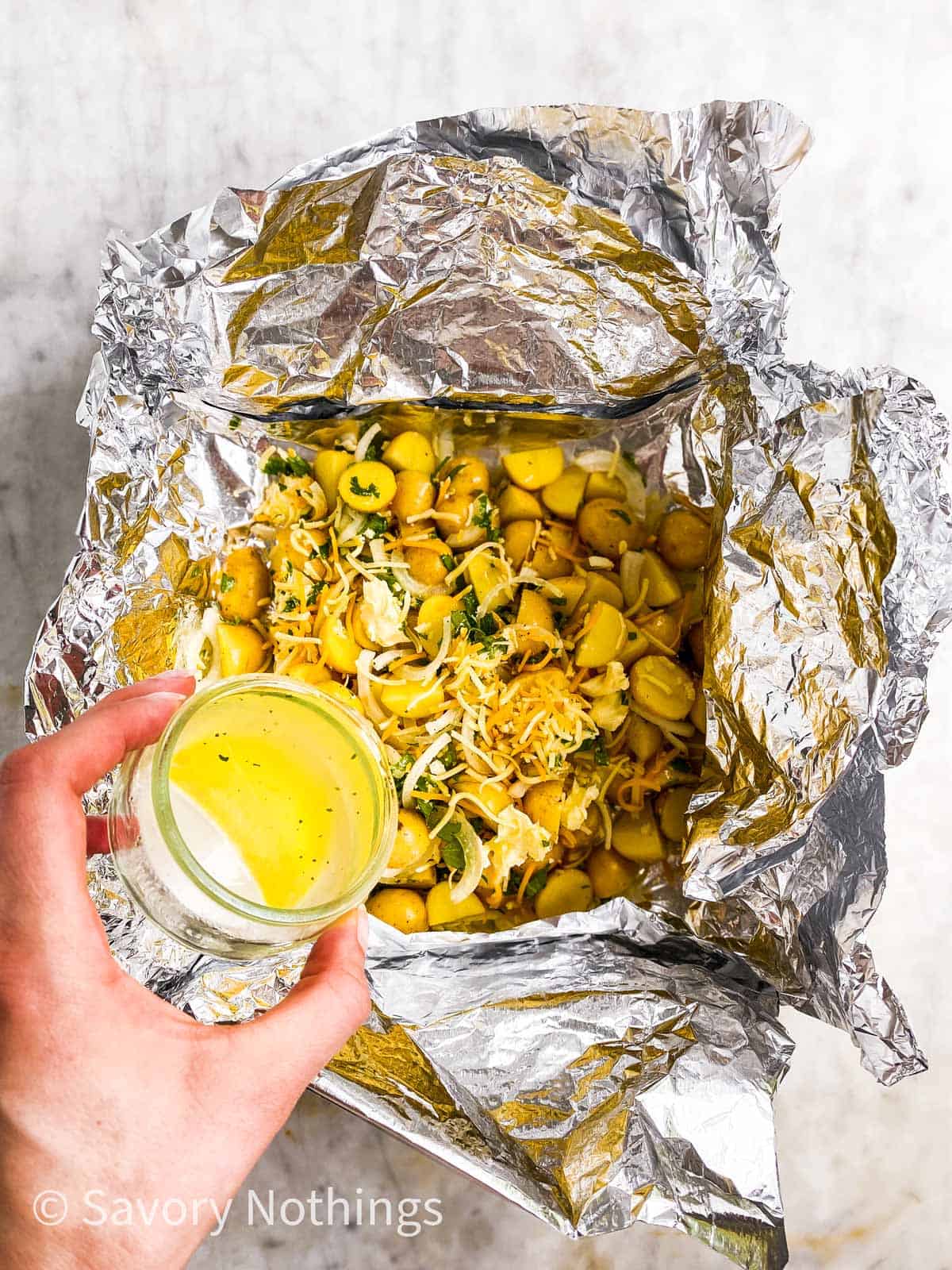 female hand pouring chicken broth over seasoned potatoes and onions in aluminium foil boat