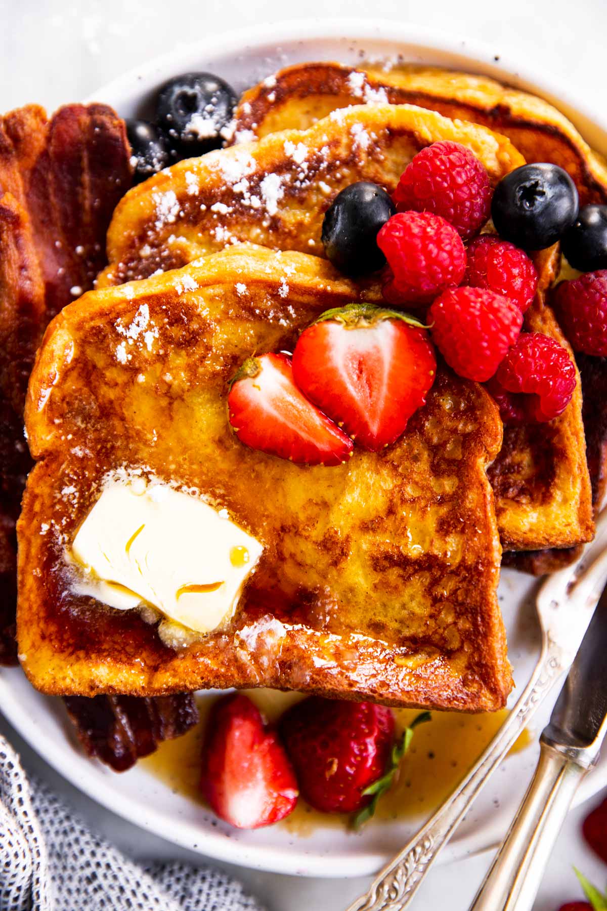 three slices of French toast on white plate with butter, bacon and berries