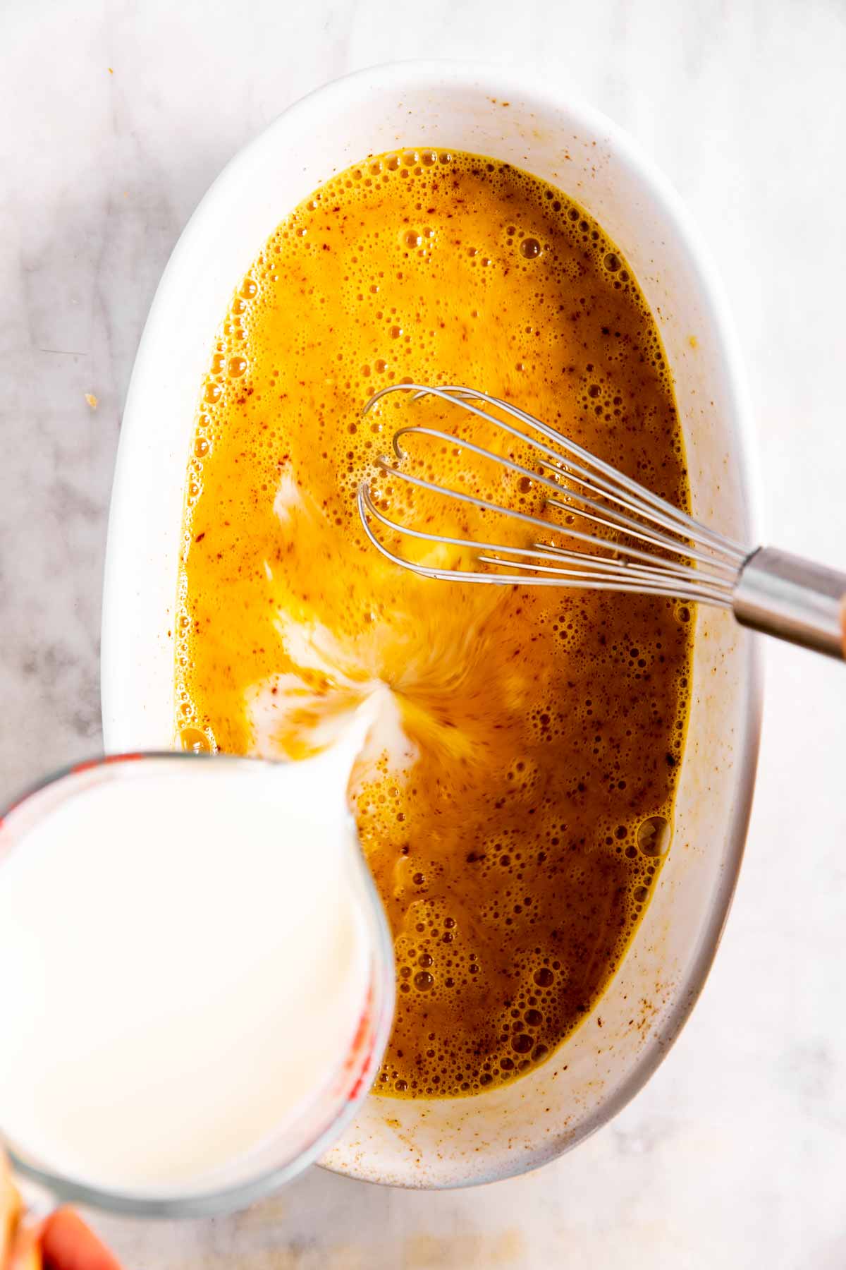 milk pouring from measuring jug into egg mixture in white dish with metal whisk