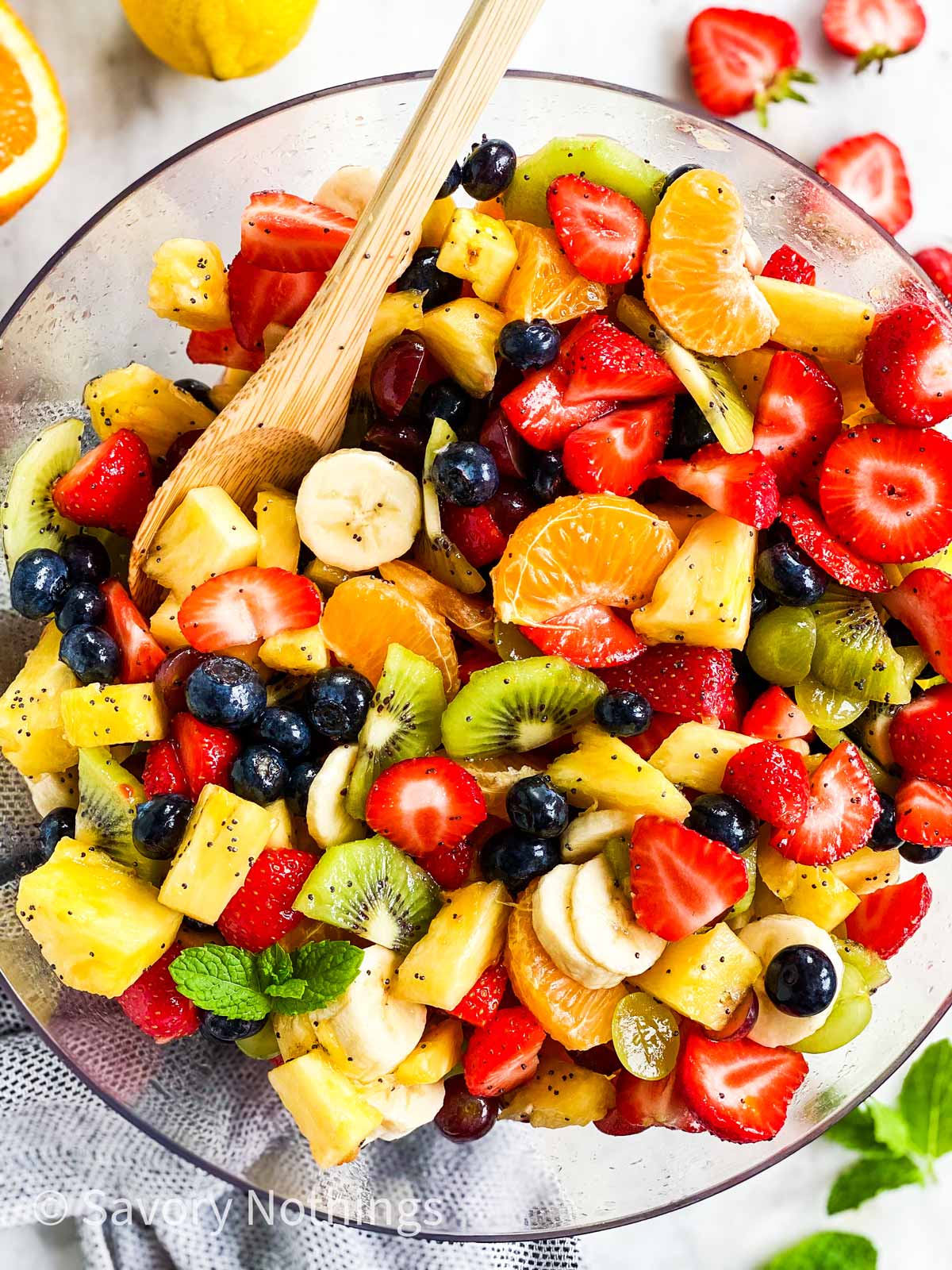 overhead view of fresh fruit salad in glass bowl with wooden spoon, surrounded by more fresh fruit