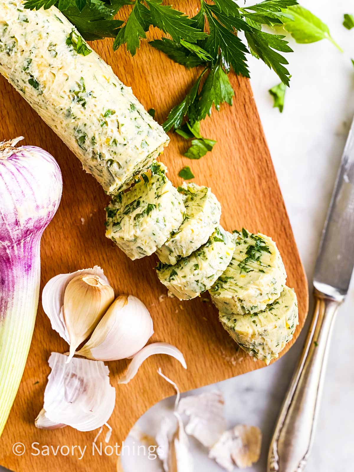 sliced garlic herb butter on wooden board with fresh garlic, parsley and a silver knife