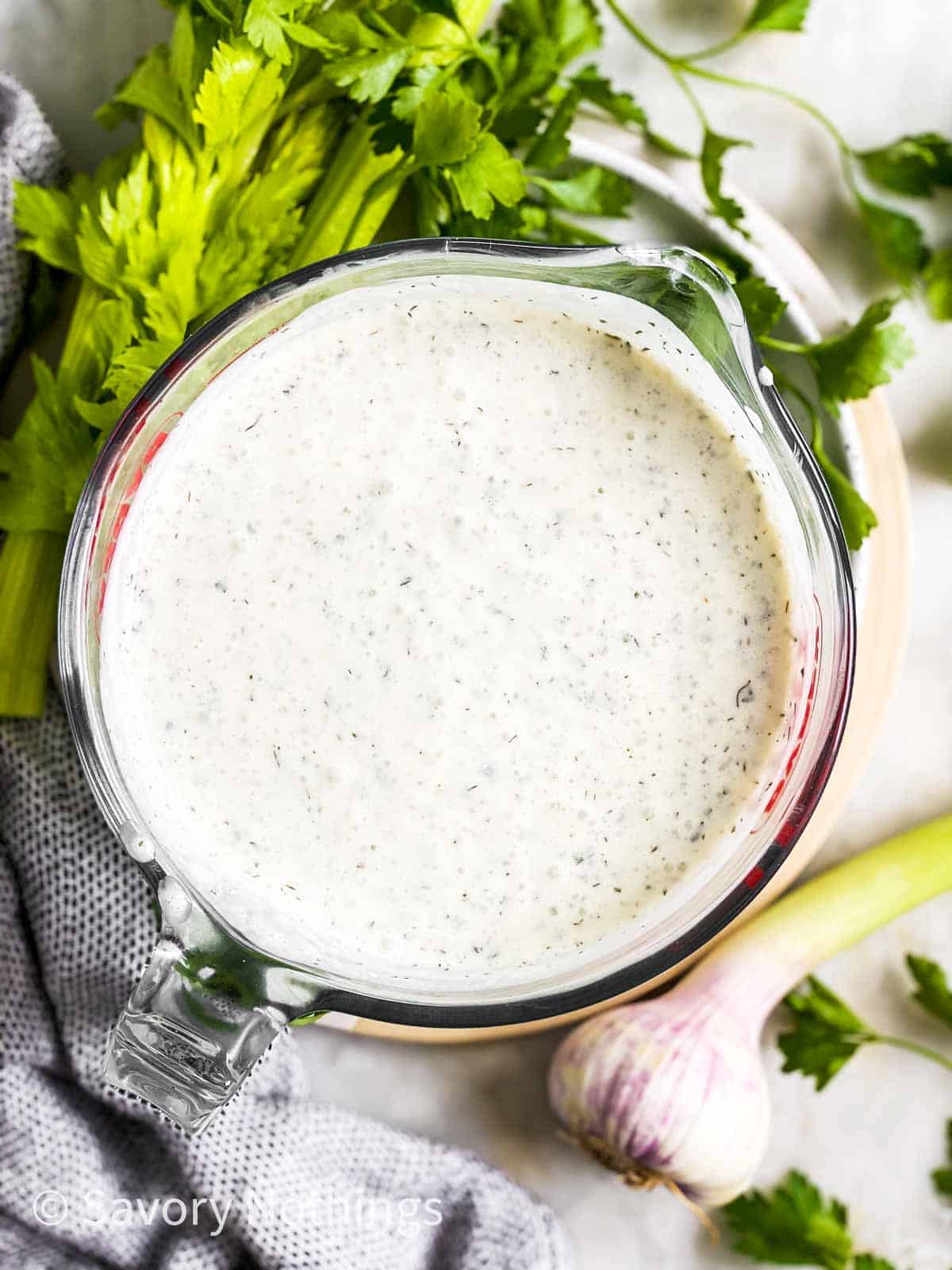 overhead view of ranch salad dressing in glass measuring jug on platter with fresh herbs and garlic