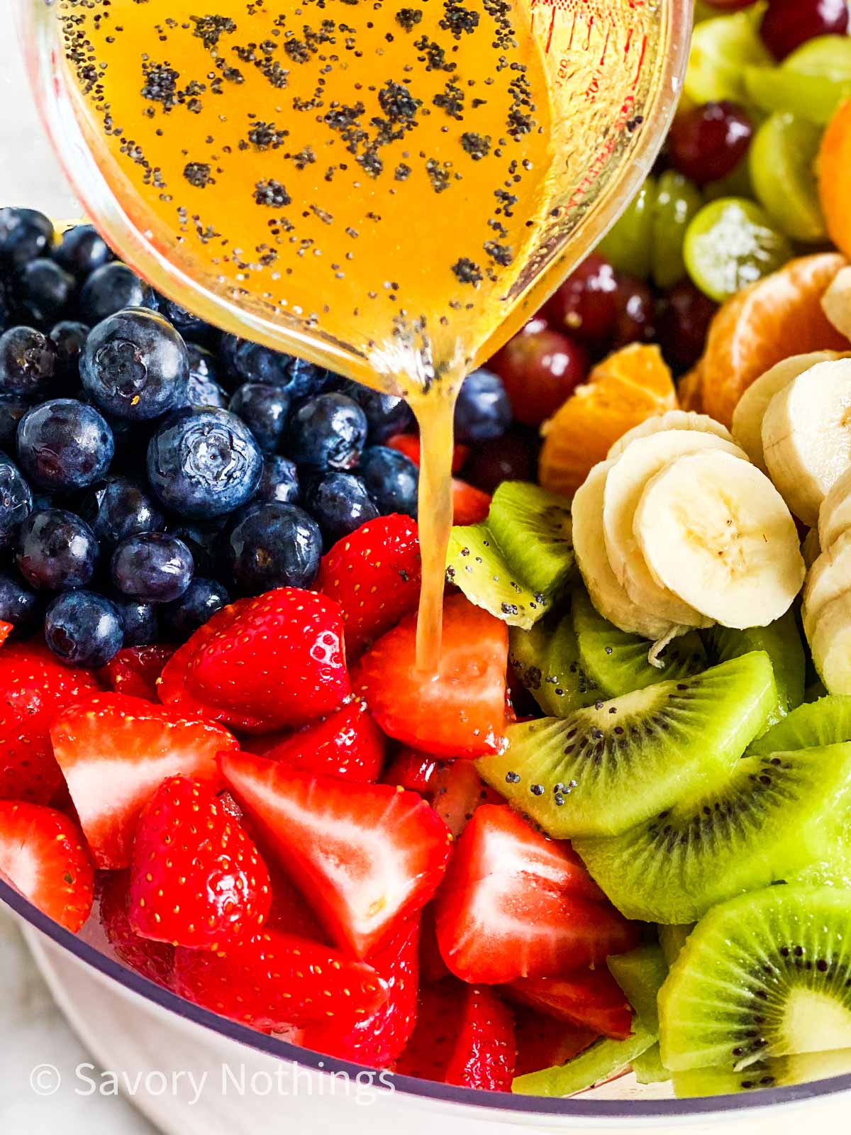 poppy seed fruit salad dressing pouring over freshly cut fruit from small glass measuring jug