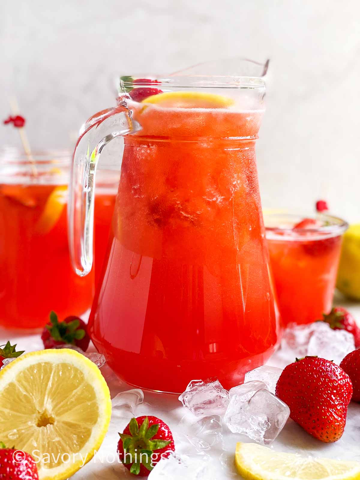 glass jug filled with strawberry lemonade surrounded by ice, strawberries, lemon and glasses full of strawberry lemonade