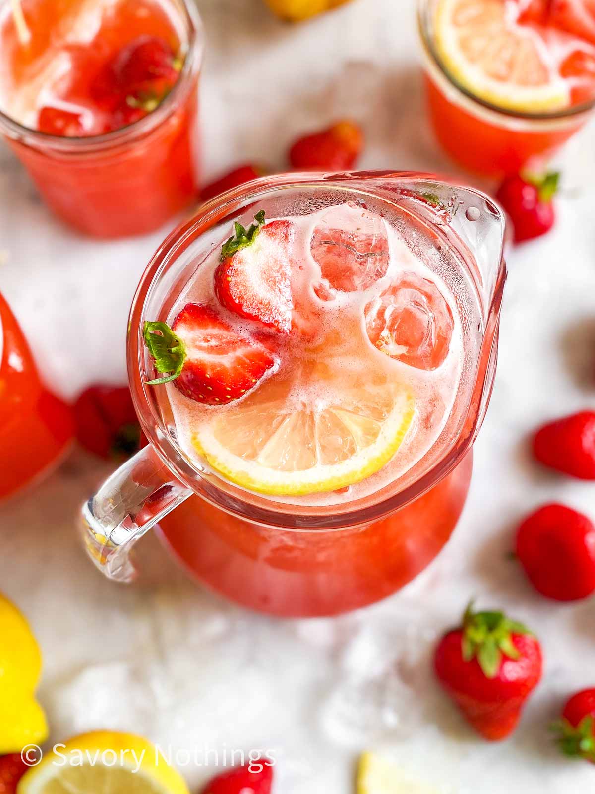 angled overhead view of glass jug filled with strawberry lemonade, lemon slices, ice cubes and sliced strawberries