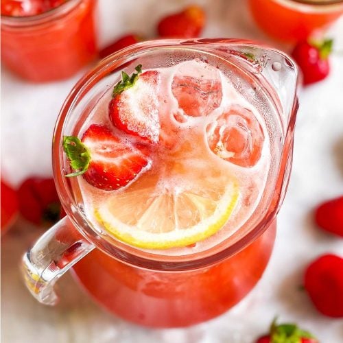 angled overhead view of glass jug filled with strawberry lemonade, lemon slices, ice cubes and sliced strawberries