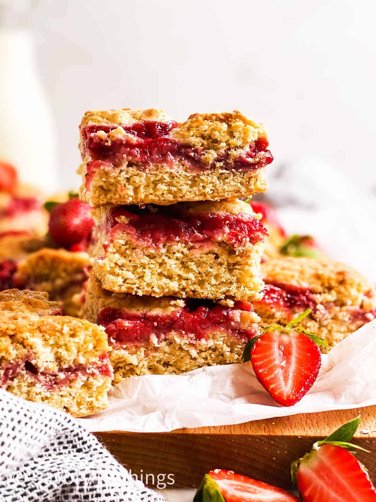 stack of three strawberry oatmeal bars on wooden board line with white baking parchment, surrounded by fresh strawberries