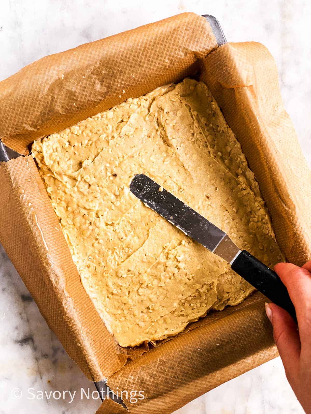 female hand using offset spatula to spread oatmeal cookie dough in lined pan