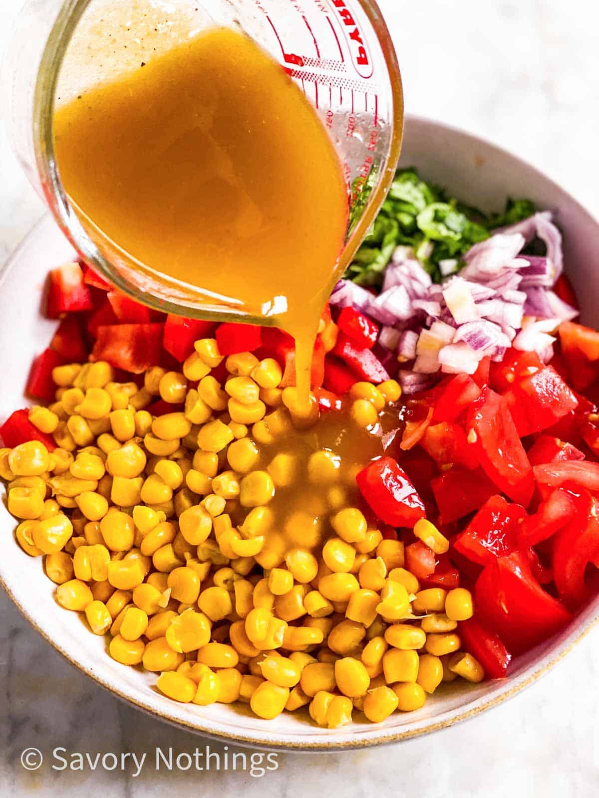 salad dressing pouring from glass measuring jug over corn salad ingredients in white bowl