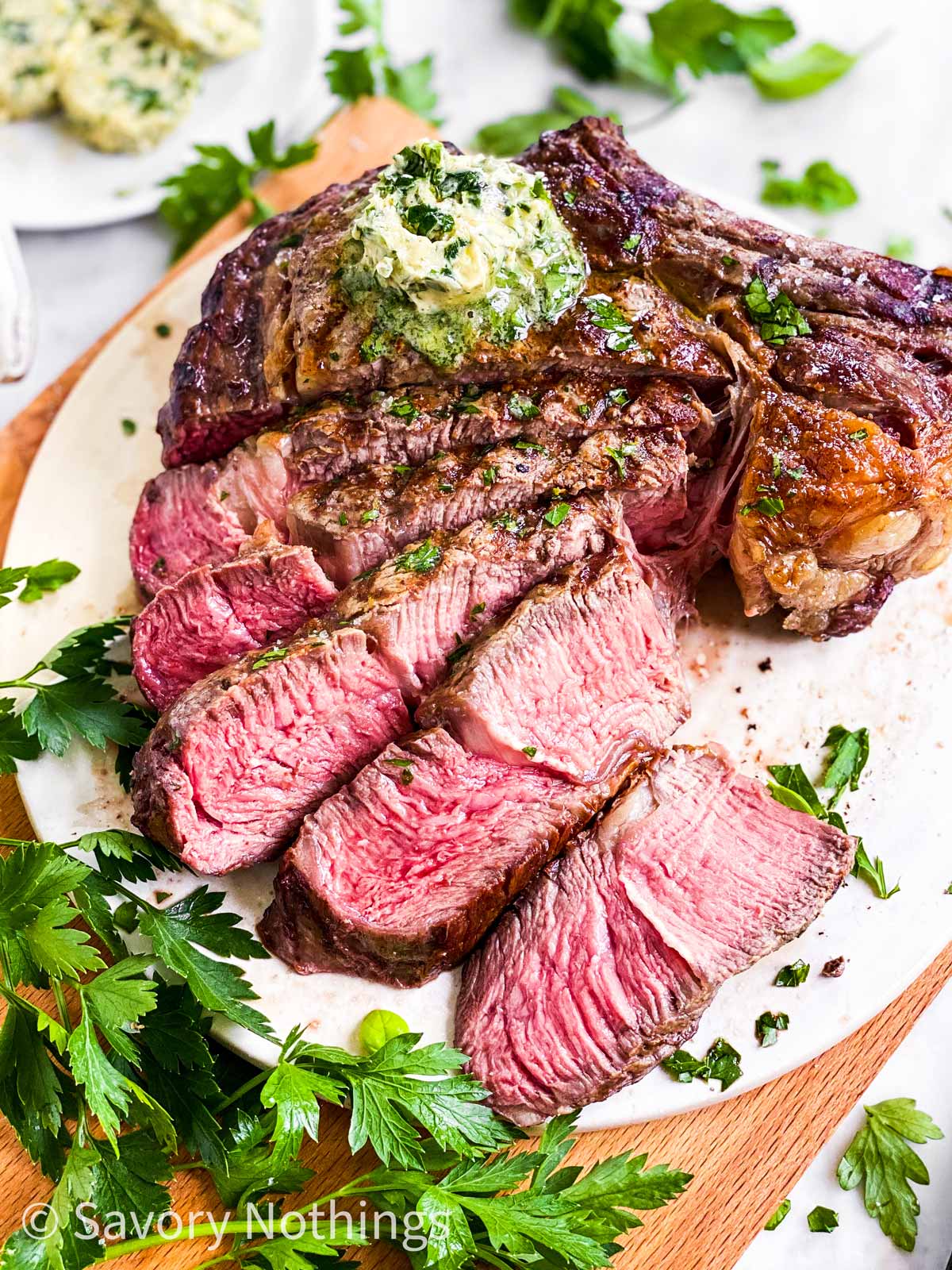 frontal view of sliced tomahawk steal on white platter with fresh parsley and garlic butter