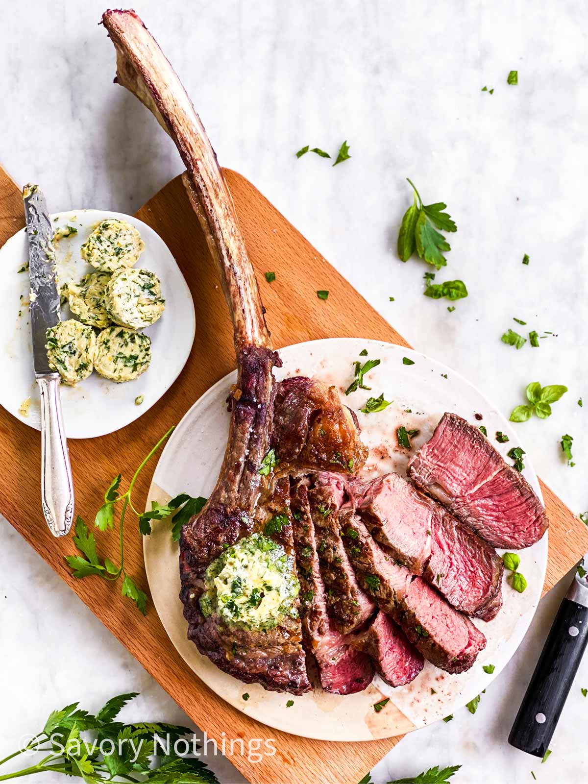 overhead view of sliced tomahawk steak on wooden board with garlic herb butter and fresh parsley