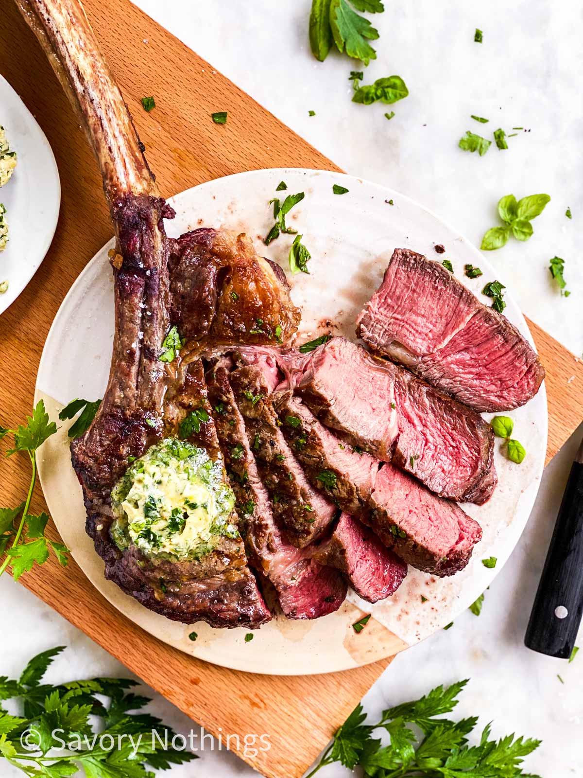sliced tomahawk steak garnished with garlic butter and fresh parsley on white platter sitting on wooden board