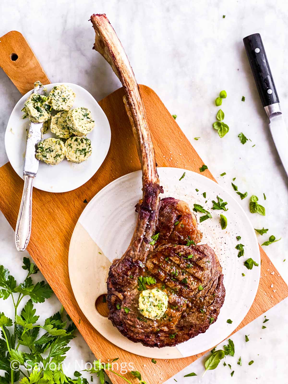 grille tomahawk steak on white platter sitting on wooden board, surrounded by steak knife, fresh parsley and garlic butter