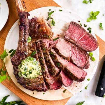 sliced tomahawk steak garnished with garlic butter and fresh parsley on white platter sitting on wooden board