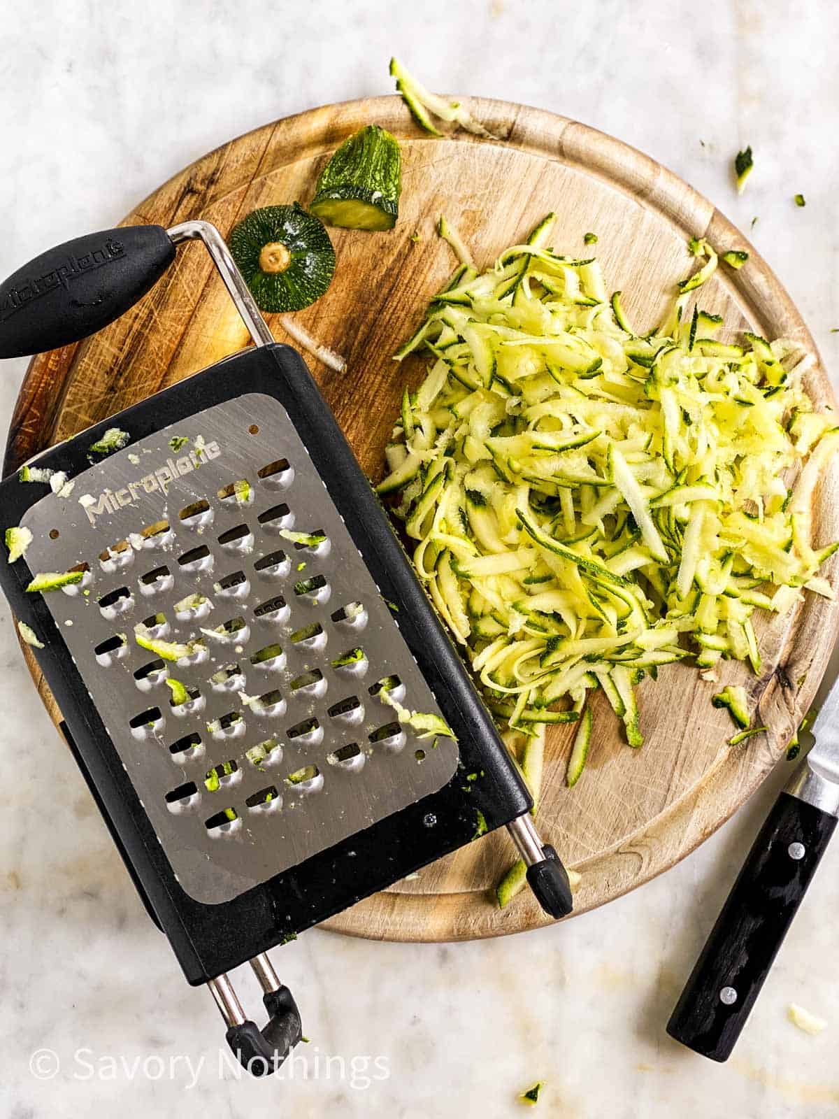 shredded zucchini on round wooden board with box grater and kitchen knife