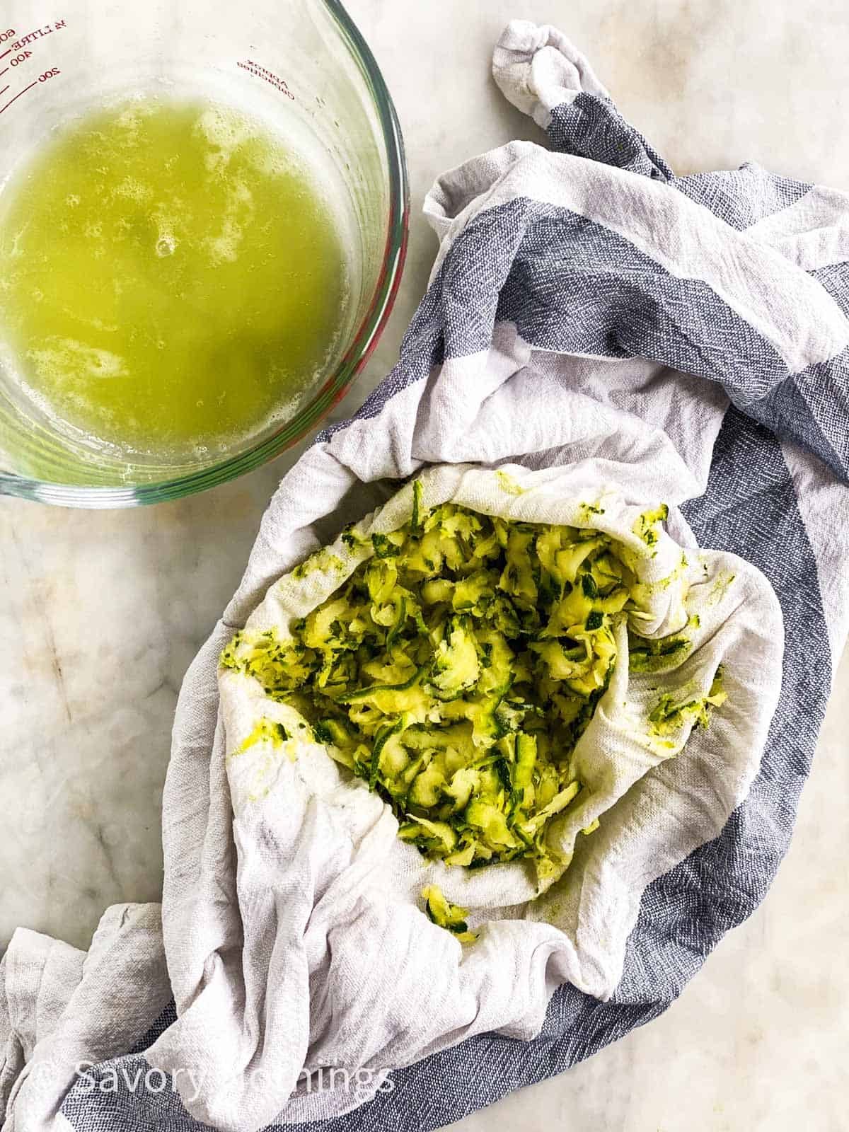 shredded and squeezed zucchini nestled in middle of blue and white kitchen towel next to glass measuring jug with zucchini juices