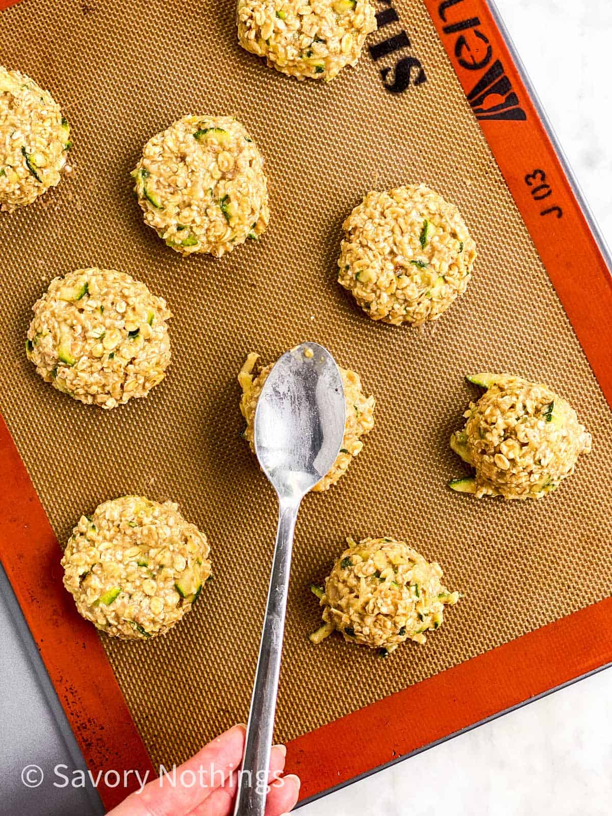 female hand using tablespoon to flatten zucchini breakfast cookies on silicone mat lined cookie sheet