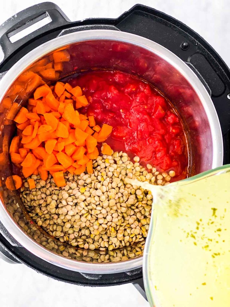 Broth pouring from glass measuring jug over lentils, diced carrots and diced tomatoes in instant pot