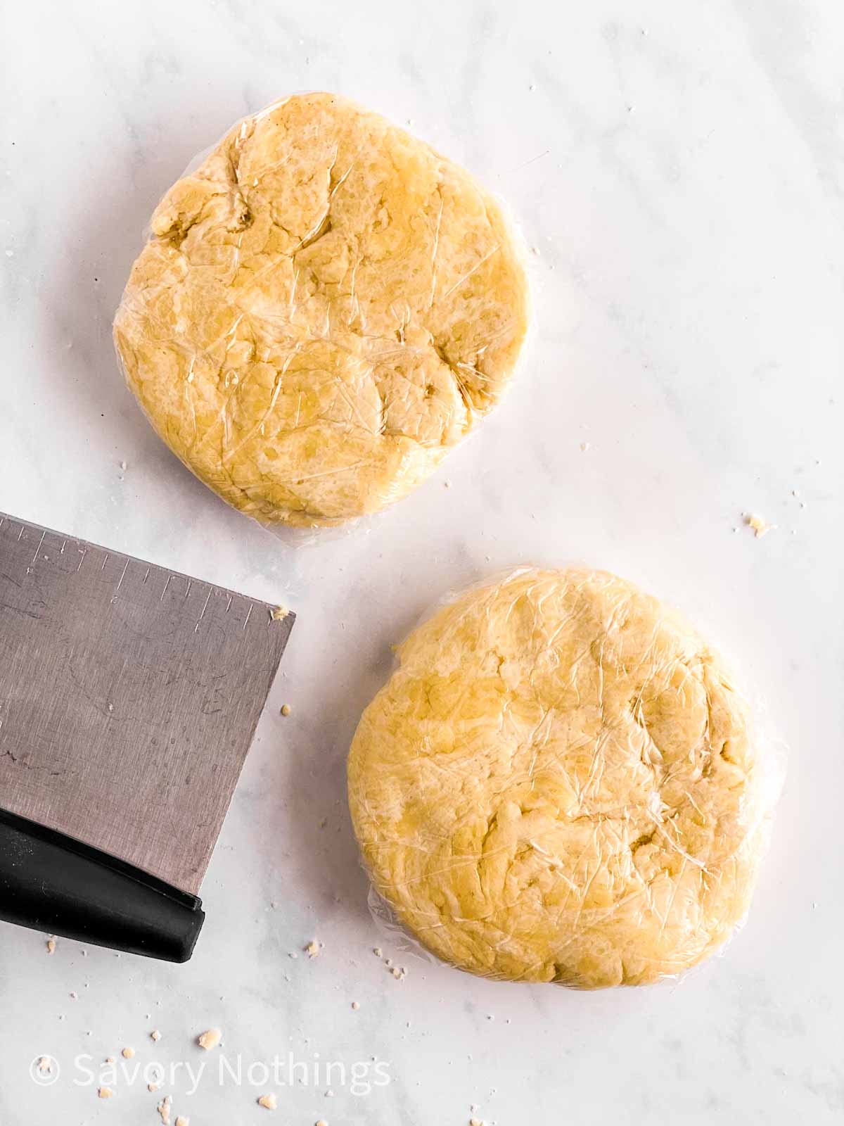 two disks of pie crust wrapped in plastic wrap next to dough scraper
