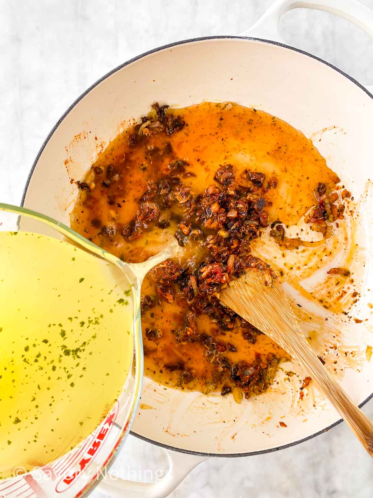 chicken broth pouring from glass measuring jug into white skillet with sautéed aromatics