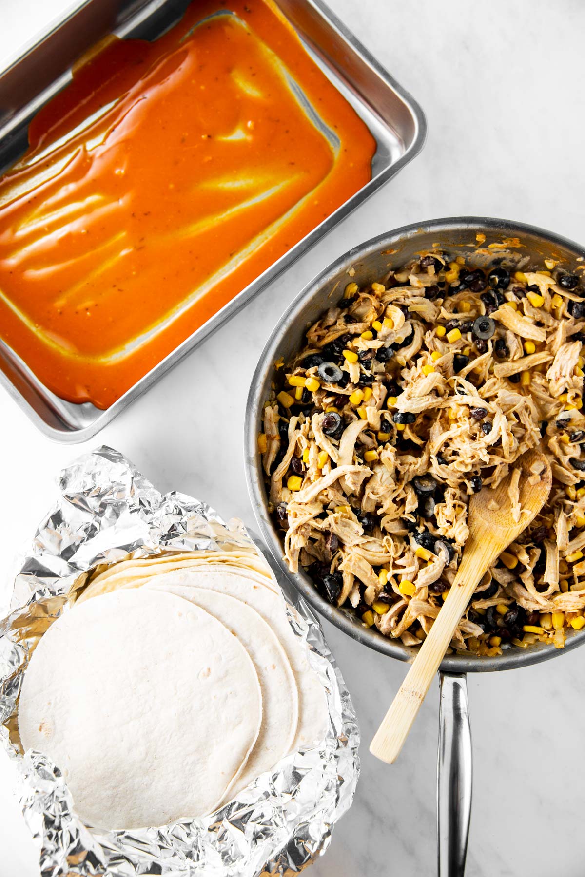 turkey enchilada filling, corn tortillas in foil and casserole dish with enchilada sauce on white surface