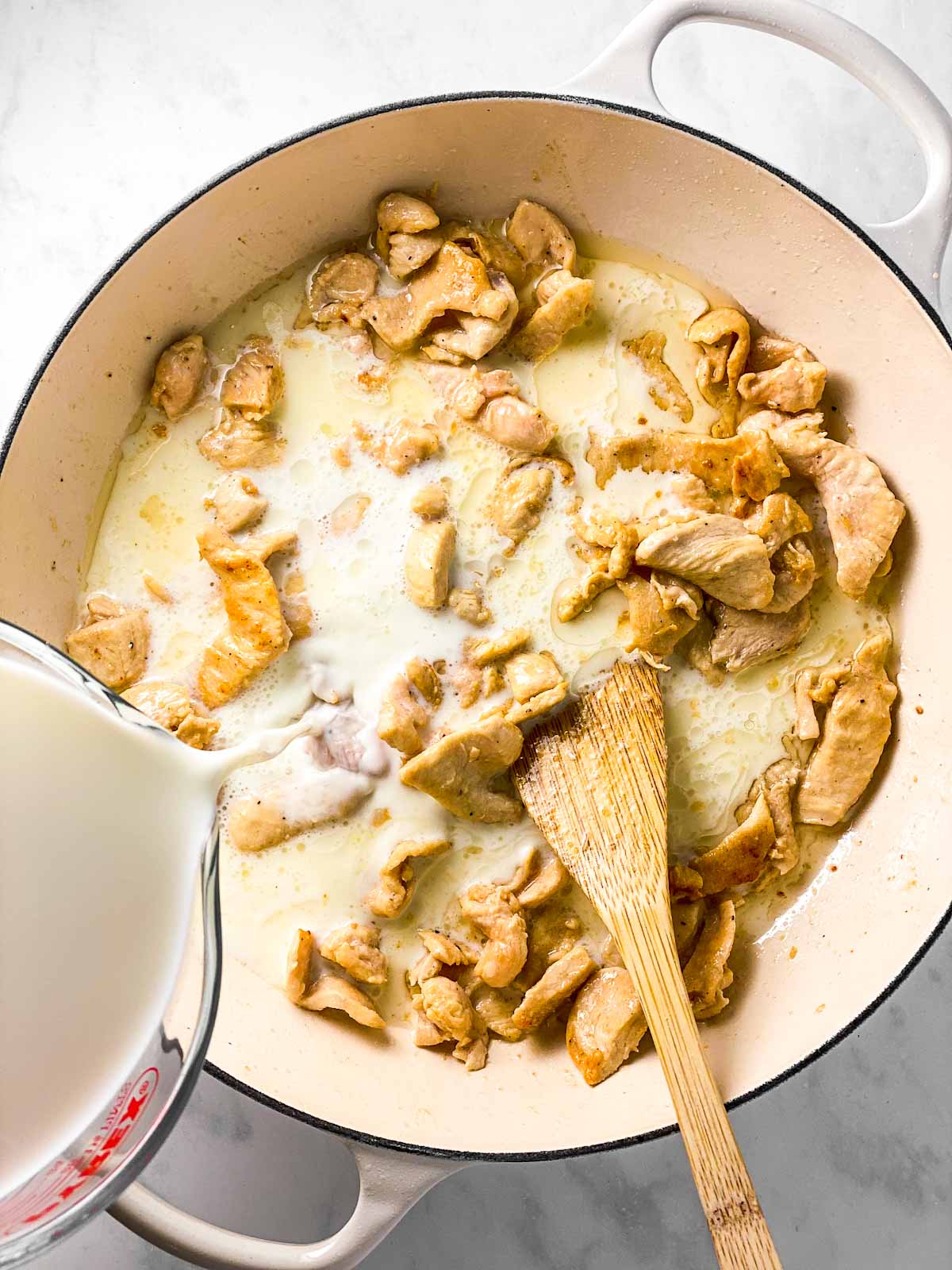 milk pouring from glass measuring jug into skillet with chicken