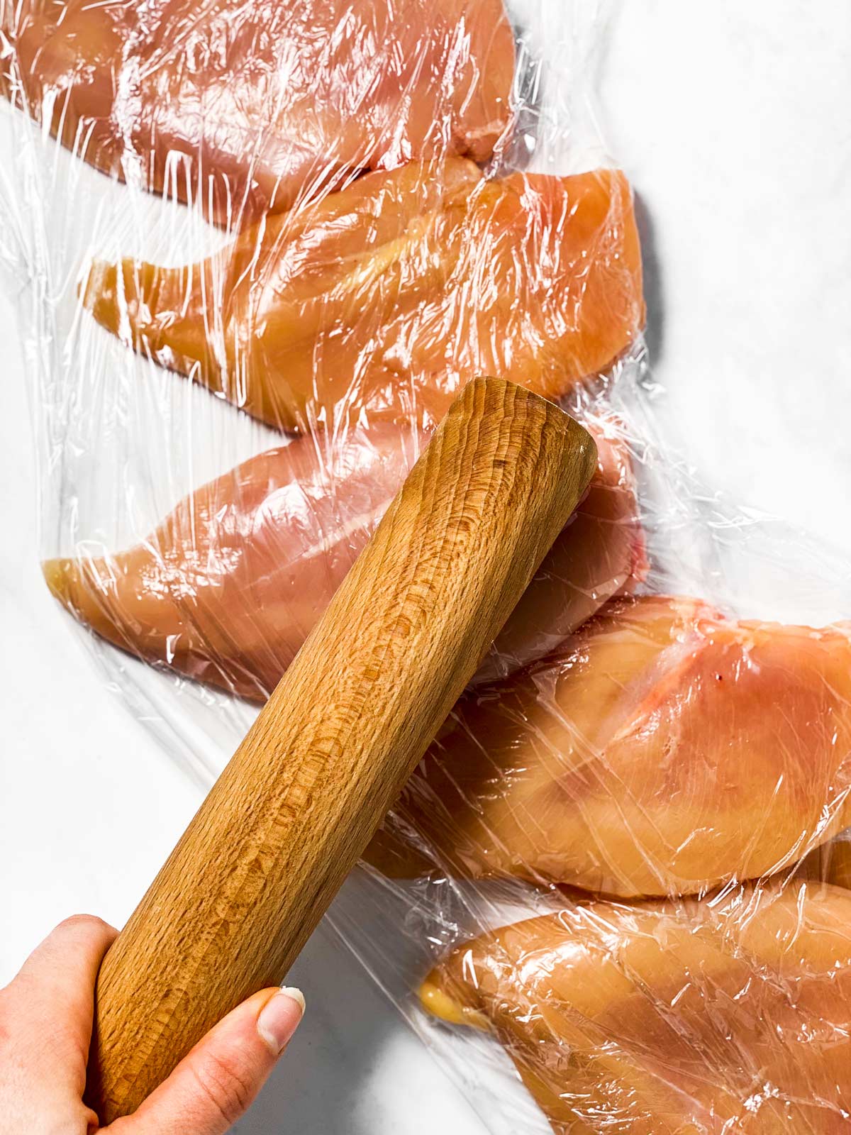 female hand using rolling pin to pound chicken breasts between plastic wrap