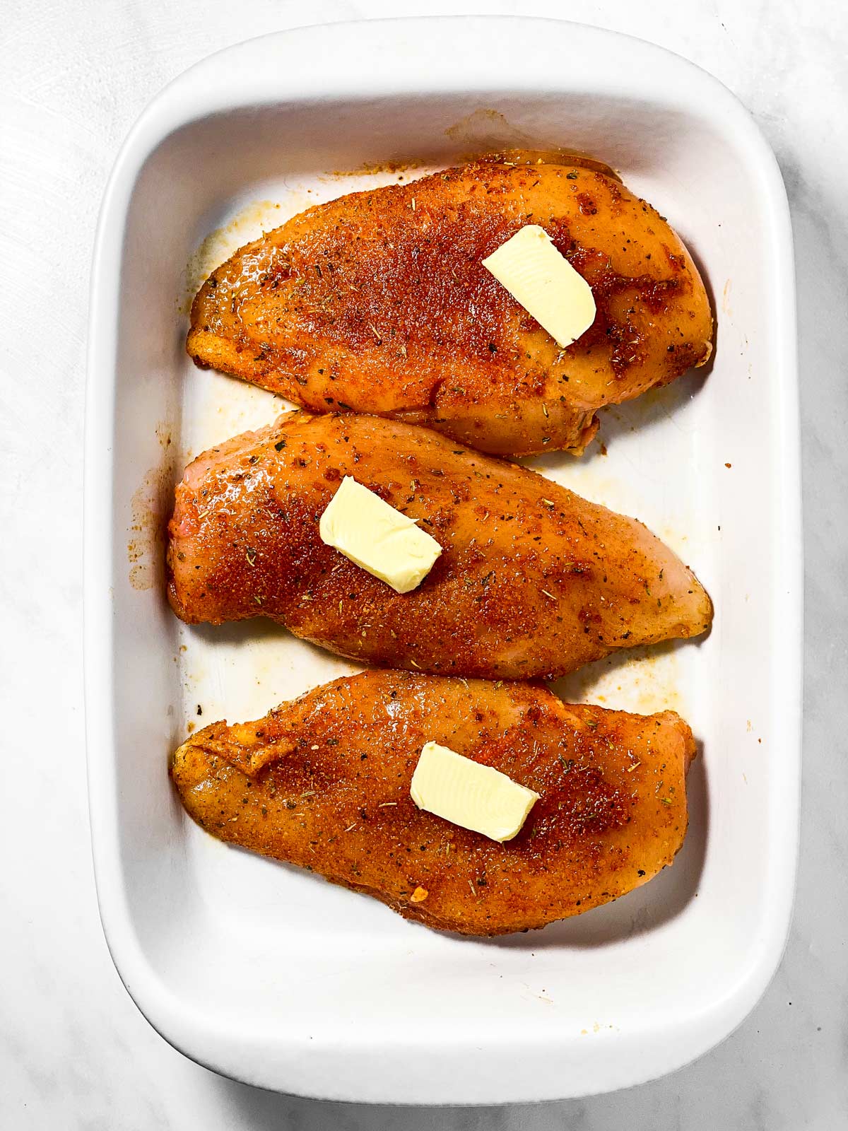 three seasoned, unbaked chicken breasts topped with butter slices in white casserole dish