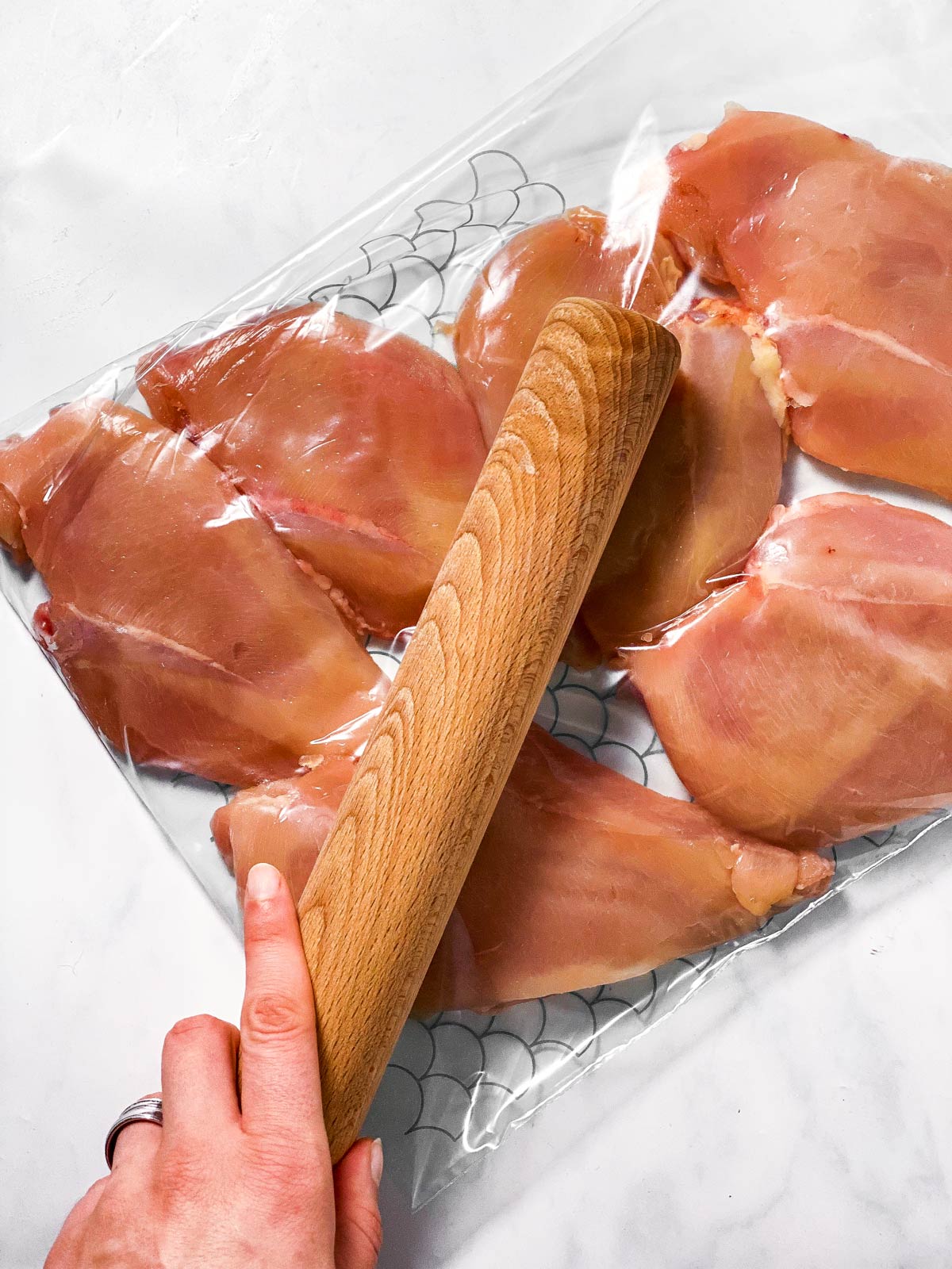 female hand using rolling pin to pound chicken in plastic bag