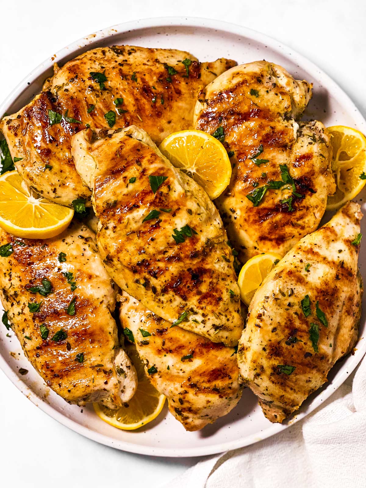 overhead view of grilled chicken breasts on white plate with chopped parsley and lemon slices
