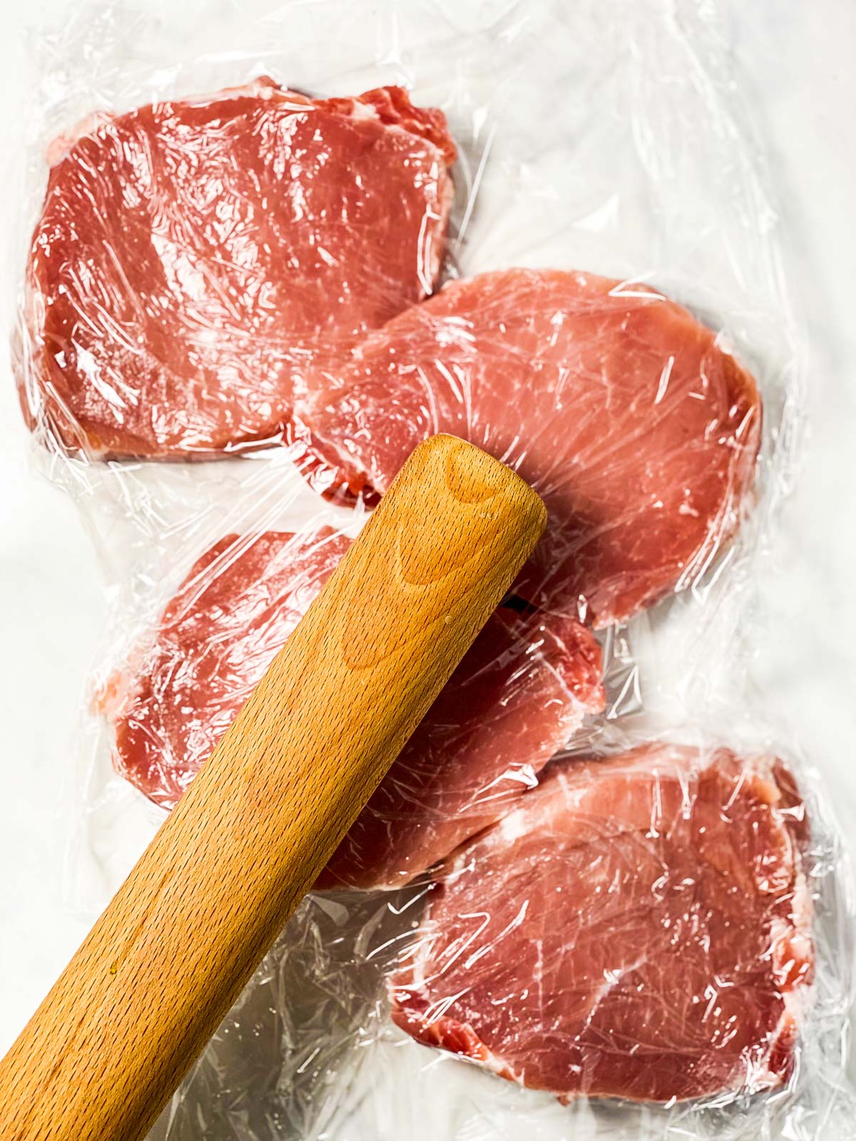 rolling pin pounding pork chops between two pieces of plastic wrap