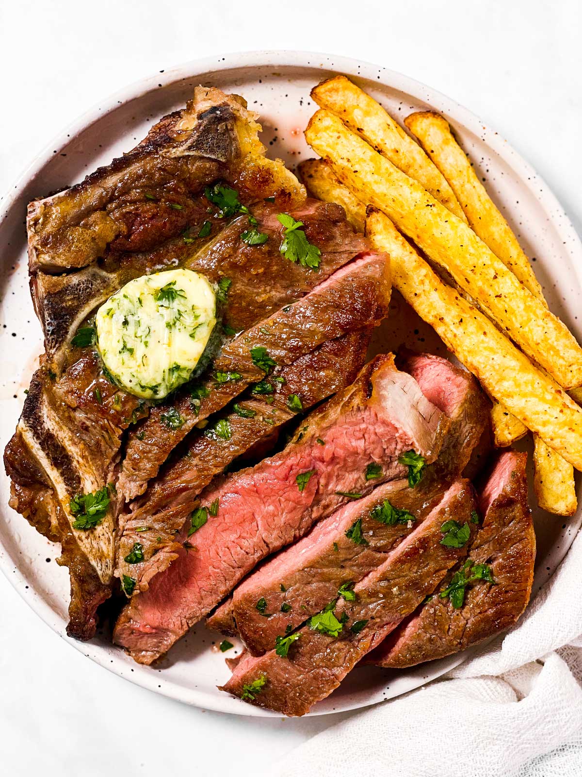 sliced steak topped with garlic butter next to french fries on white plate