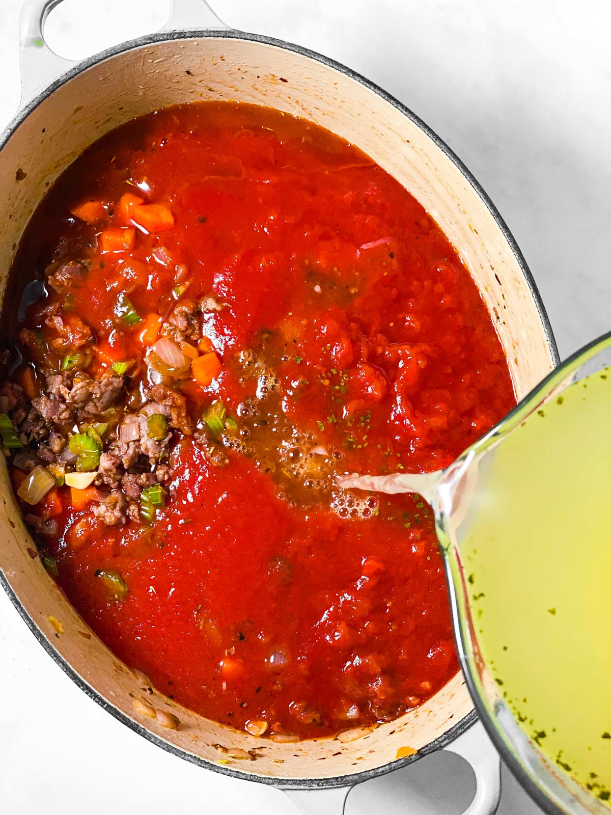 chicken broth pouring from glass measuring jug into Dutch oven with tomato sauce