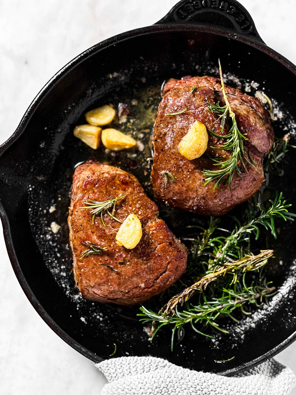 overhead view of two cooked filet mignons in black cast iron pan with garlic and rosemary